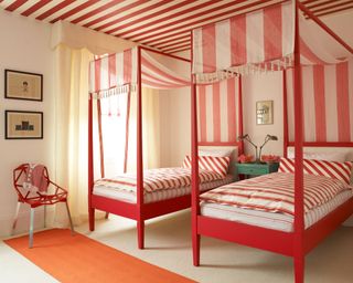 Red bedroom with two painted four poster beds, striped red and white duvet covers and sheets and matching bespoke striped canopies, turquoise side table with two headed lamp, flowers heads in bowls, framed prints, red metal geometric shaped chair.