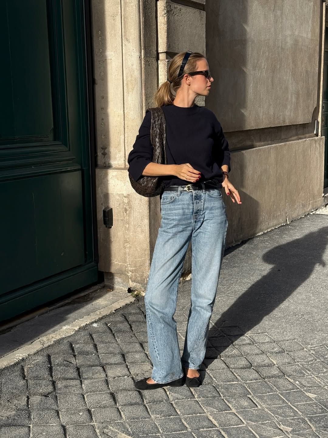 A woman wears a black sweater, blue jeans, and black ballet flats with a woven shoulder bag.