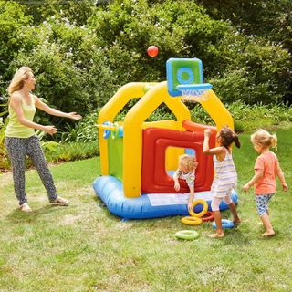 a women and three children on a grassy area playing with a red, yellow, blue and green small bouncy castle that has a basketball ring and ring toss