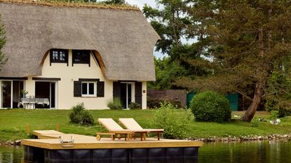 a thatched cottage on the edge of a pond in Normandy, France surrounded by greenery, tree, ducks with a deck for sunbathing