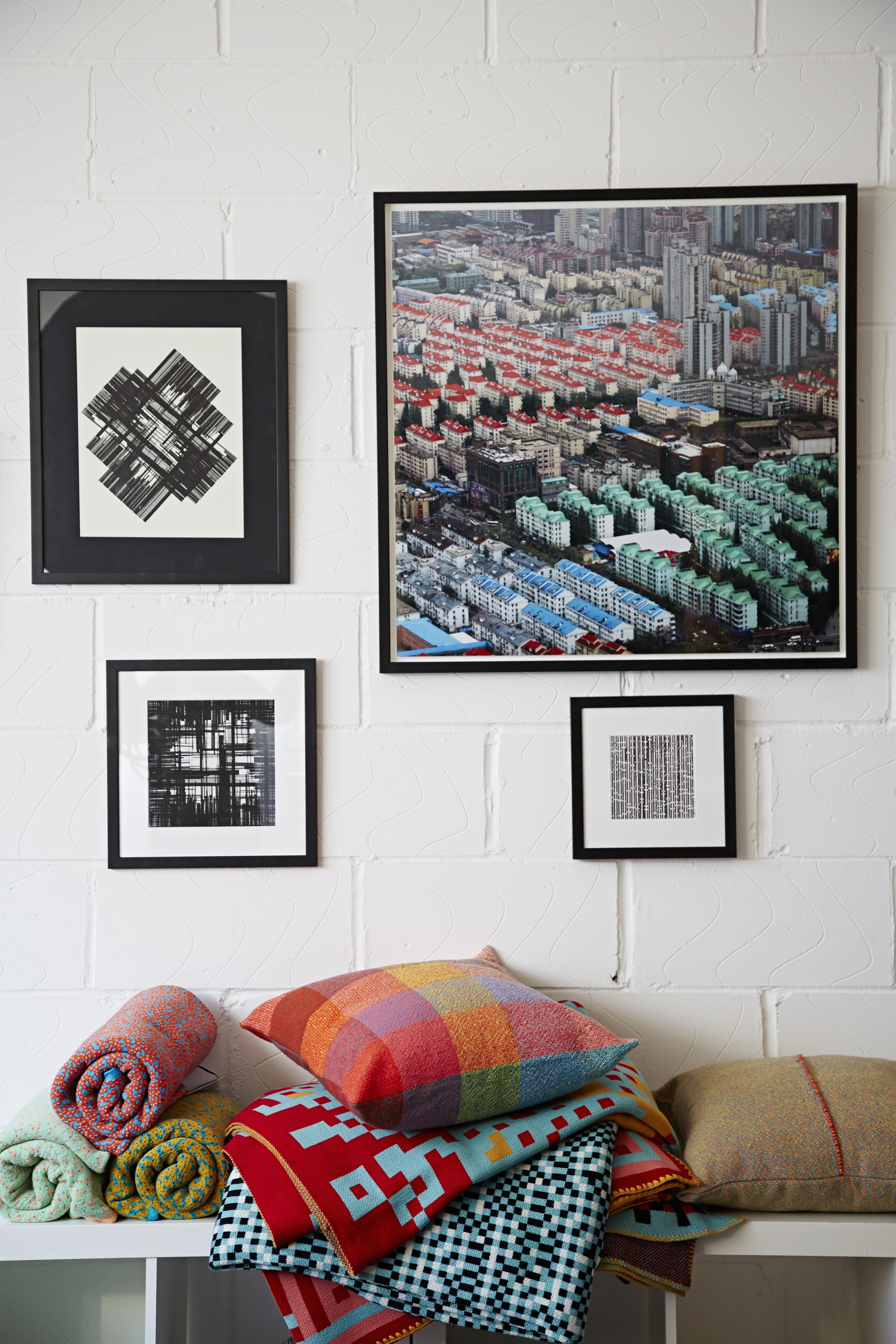 A pile of stacked and rolled colorful blankets against a white wall with artwork above