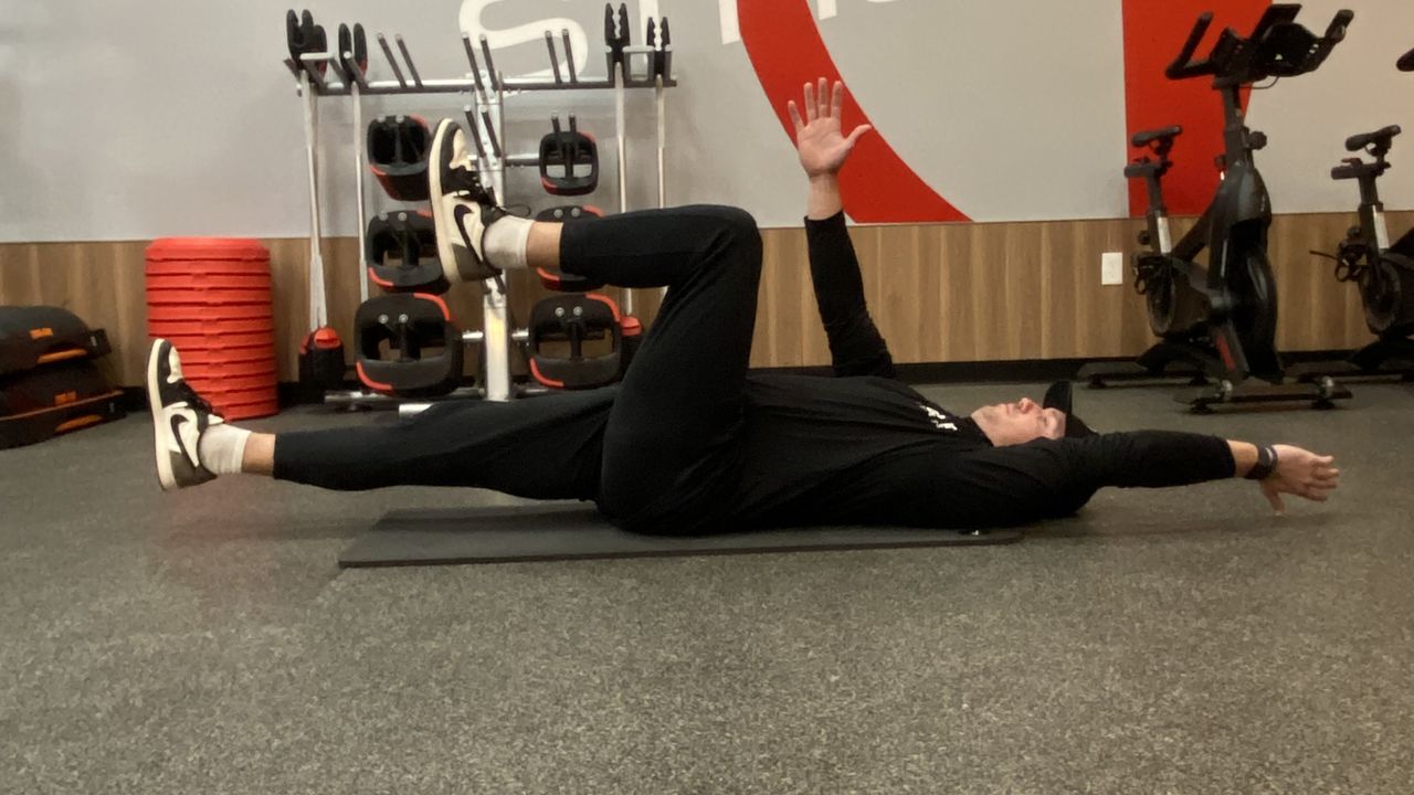 Man lies on his back on exercise mat demonstrating the dead bug exercise. He wears Nike trainers, black pants, black sweatshirt and black cap.