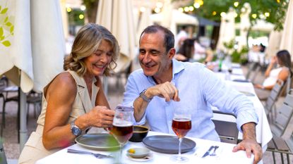 A happy retired couple is dining out, drinking wine at a nice restaurant. They are smiling and looking at her smartphone.