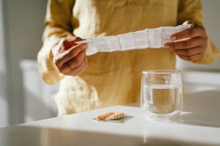 Woman standing at a table reading a small sheet of instructions from a pill box. The pills are in a sleeve on the table next to a glass of water