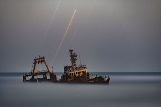 stars streak against a grey sky. an old, rusty boat sits in the foreground, shrouded in fog that covers the ground.