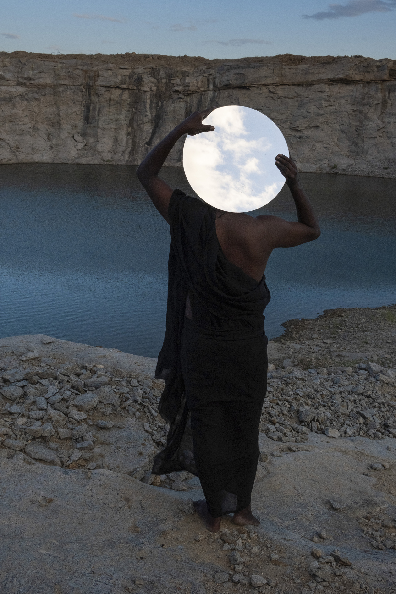 A person in a black dress stands at a rocky cliff's edge, holding a round mirror overhead that reflects a partly cloudy sky, facing a calm, blue lake