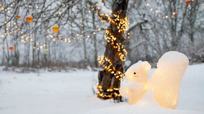 A lit up squirrel statue in the snow under a tree decorated with lights and ornaments