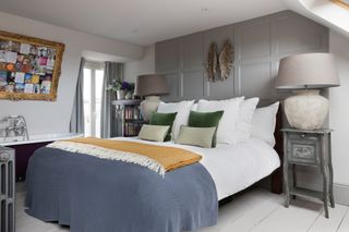 Pippa Jones house: master bedroom with white painted floorboards, grey panelled wall, bed with blue and yellow throws and distressed bedside tables with oversized lamps