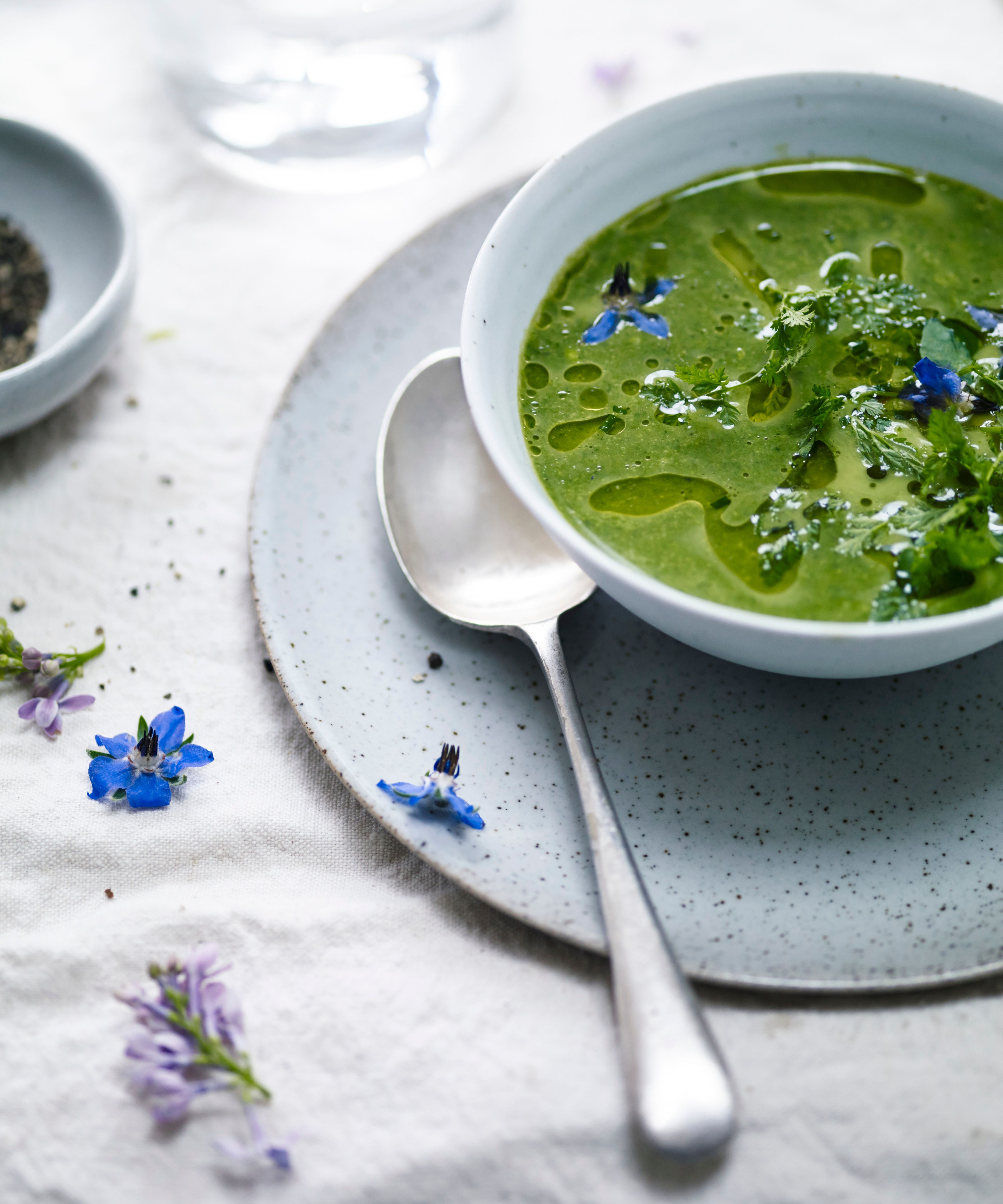 green herb soup with blue edible flowers in a speckled bowl on a plates with flowers scattered around the tablecloth