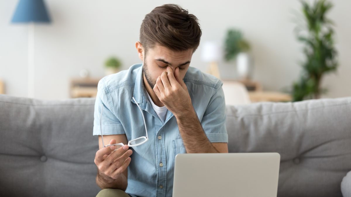 A young man sitting on a sofa in front of a laptop rubbing his eyes having taken his glasses off