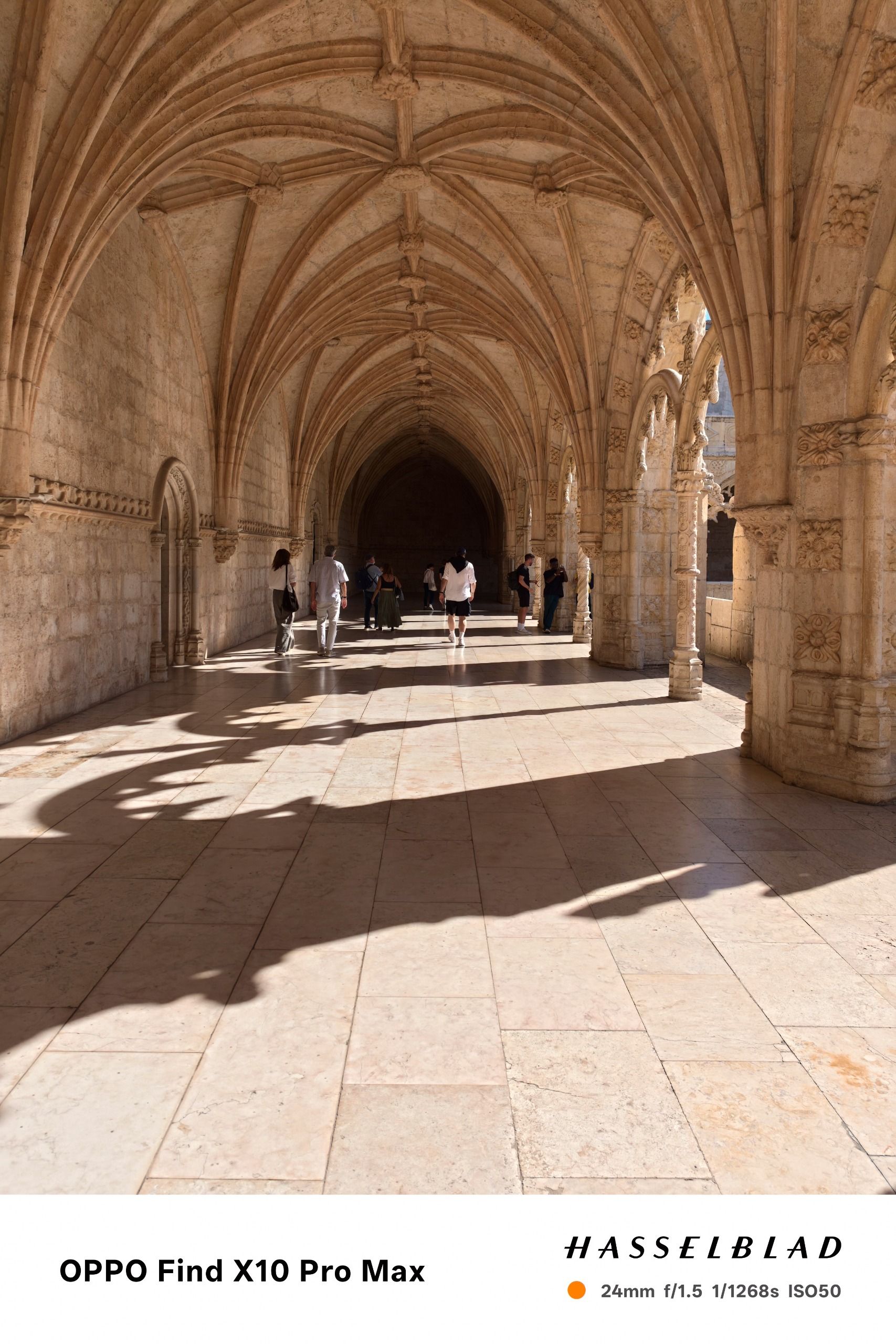 Sunlit vaulted cloister corridor with visitors walking beneath intricate stone arches.