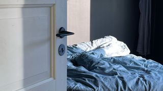 The image shows a bedroom door open and an unmade bed bathed in natural light in the background