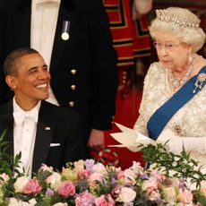 Queen Elizabeth wearing a tiara giving a speech standing up at a banquet table next to Barack Obama