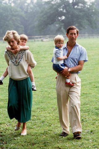 Princess Diana wearing a green skirt and white sweater giving Prince William a piggyback in grass next to Prince Charles, who is wearing a blue shirt and khakis holding Prince Harry