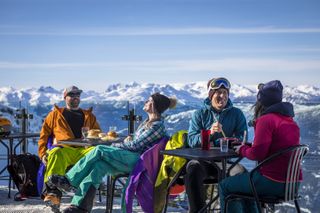 A group of people sitting at outdoor bistro tables at a ski resort with a snow-capped mountain range on the horizon