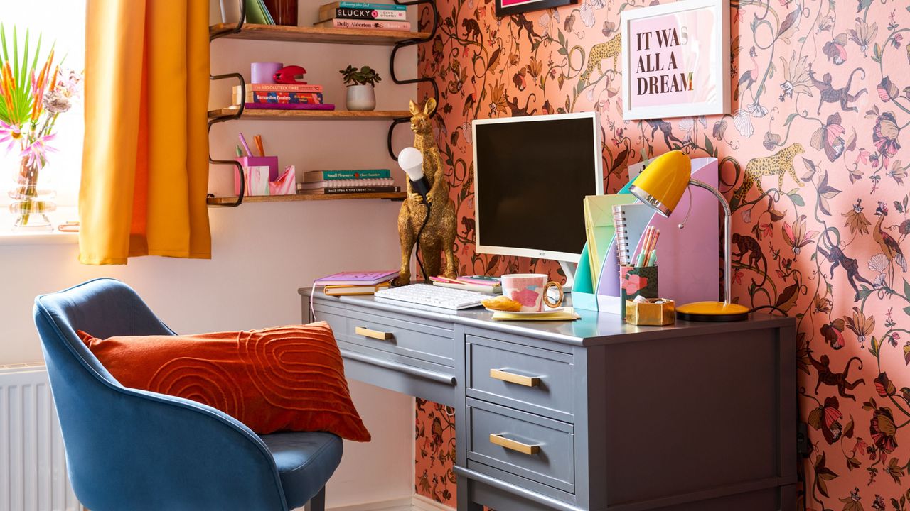 Narrow office ideas are useful to know. Here is a colorful home office with yellow curtains, wooden wall shelf with decor, pink patterned wallpaper with wall art, a navy blue office chair, a dark gray desk