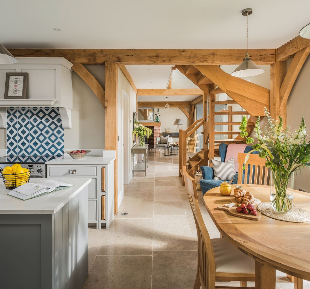 An open-plan kitchen-diner with oak beams, a wooden dinner table, a blue armchair and an oak staircase