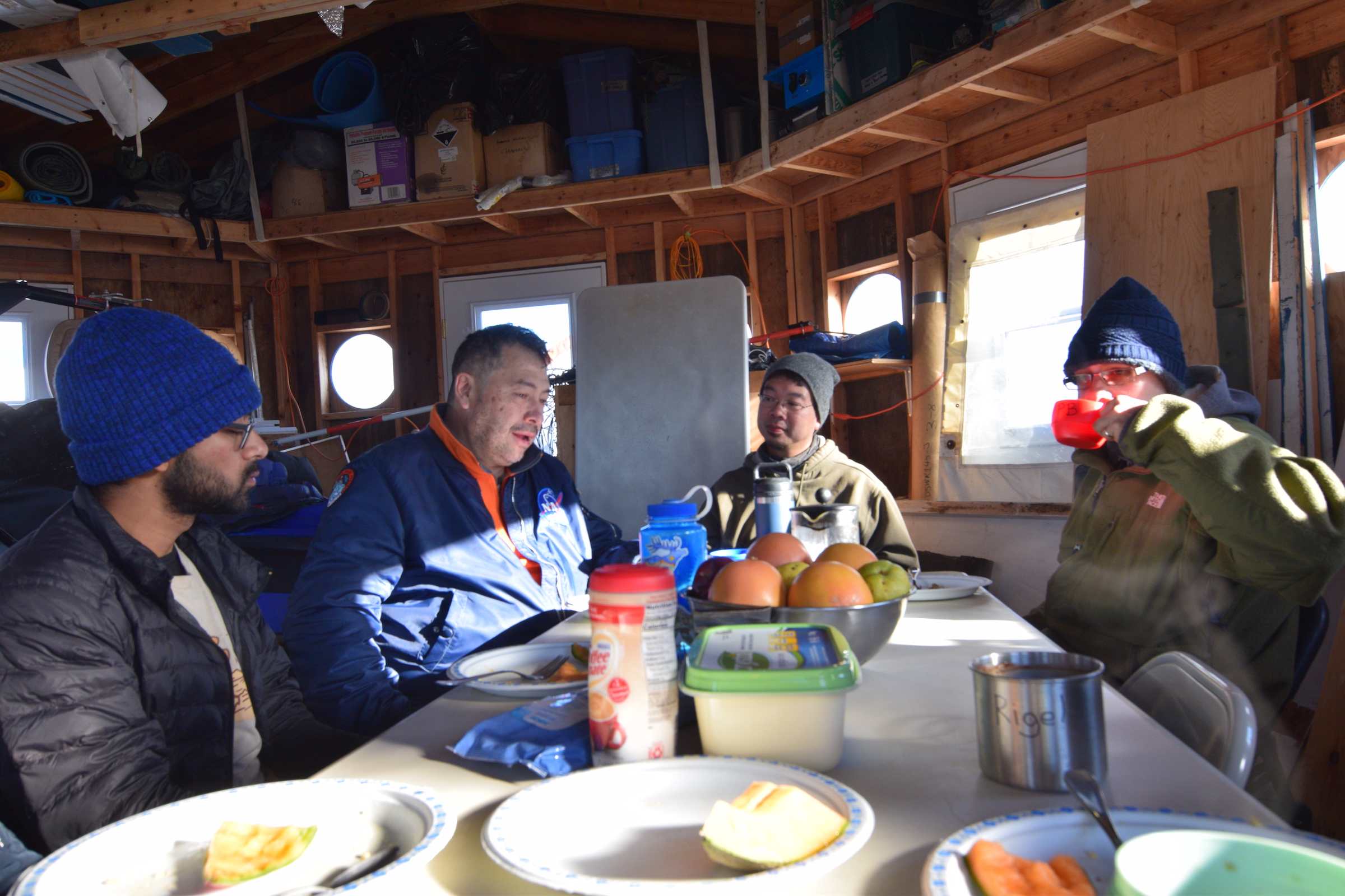 Men in coats and winter hats play cards inside an Arctic base camp structure.