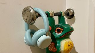 CMF Headphone Pro in pistachio green held in a hand or hung on a barbell, being pushed up by a carved frog 