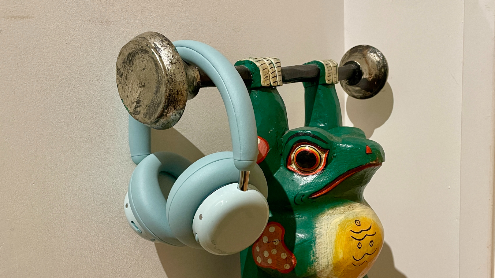 CMF Headphone Pro in pistachio green held in a hand or hung on a barbell, being pushed up by a carved frog 