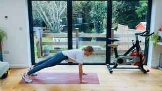 Maddy Biddulph performs a high plank on a yoga mat in her living room. She wears leggings, a t-shirt and sneakers. She is on the ground, with her body held straight and only her hands and toes touching the floor. Her arms are also straight and her head is facing down. Behind her we see an exercise bike and glass sliding doors leading to a garden.