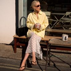 British influencer Chloe Butler sitting on a bench outside of a London cafe holding a mug while wearing oval sunglasses, a butter yellow button-down shirt, sheer white lace maxi skirt, a sleek shoulder bag, and brown flat flip-flop sandals