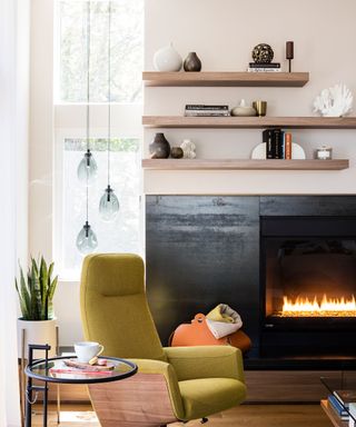 Floating wooden shelves minimally styled in modern living room with fabric chair, glass side table and black painted fireplace