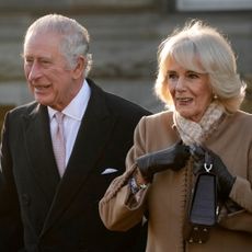 King Charles wears a black coat and white shirt while Queen Camilla wears a camel coat with black leather gloves