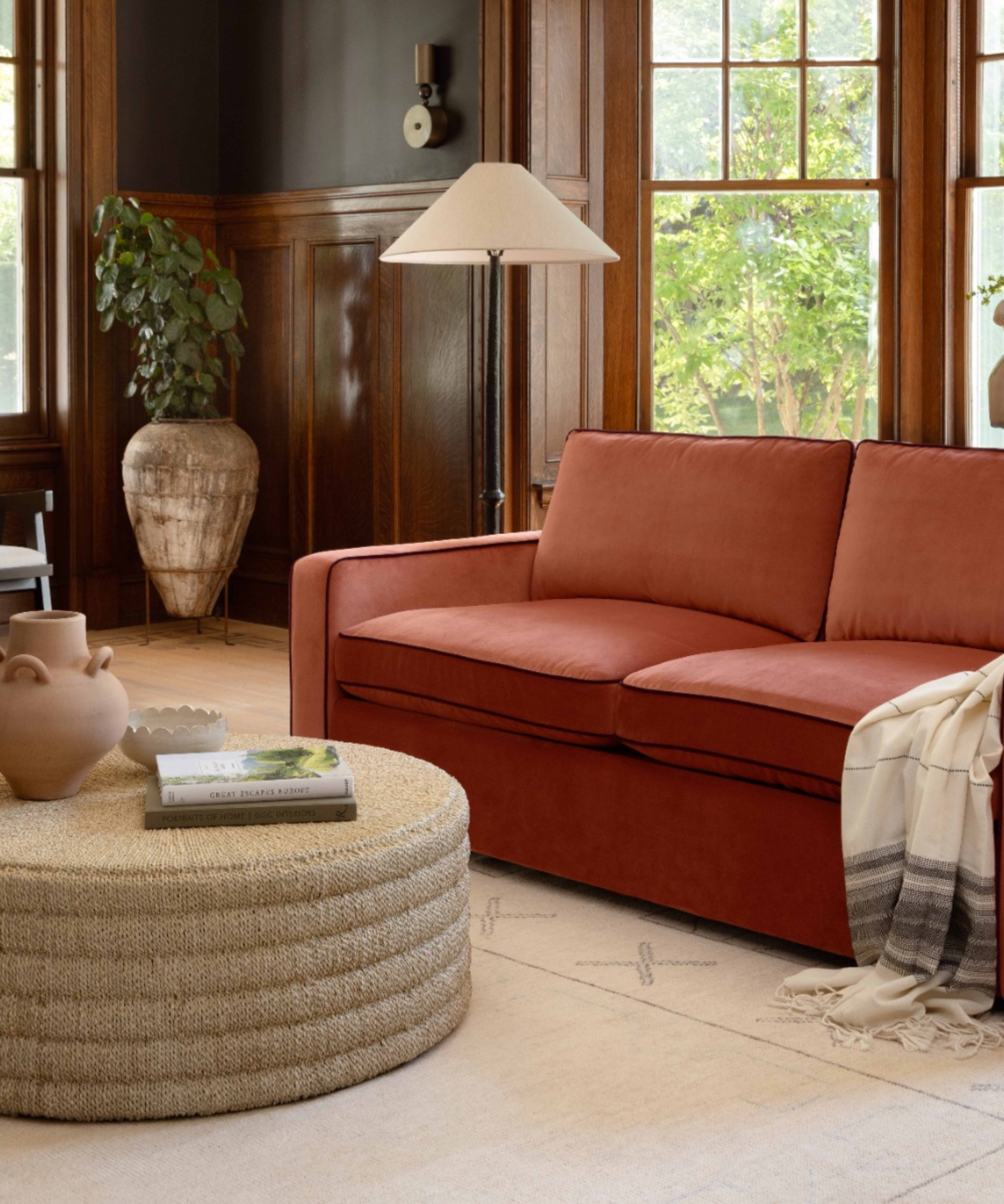 Piped-edge terracotta sleeper sofa styled in a neutral living room with decorative wooden paneling, a woven coffee table, and large bay windows.