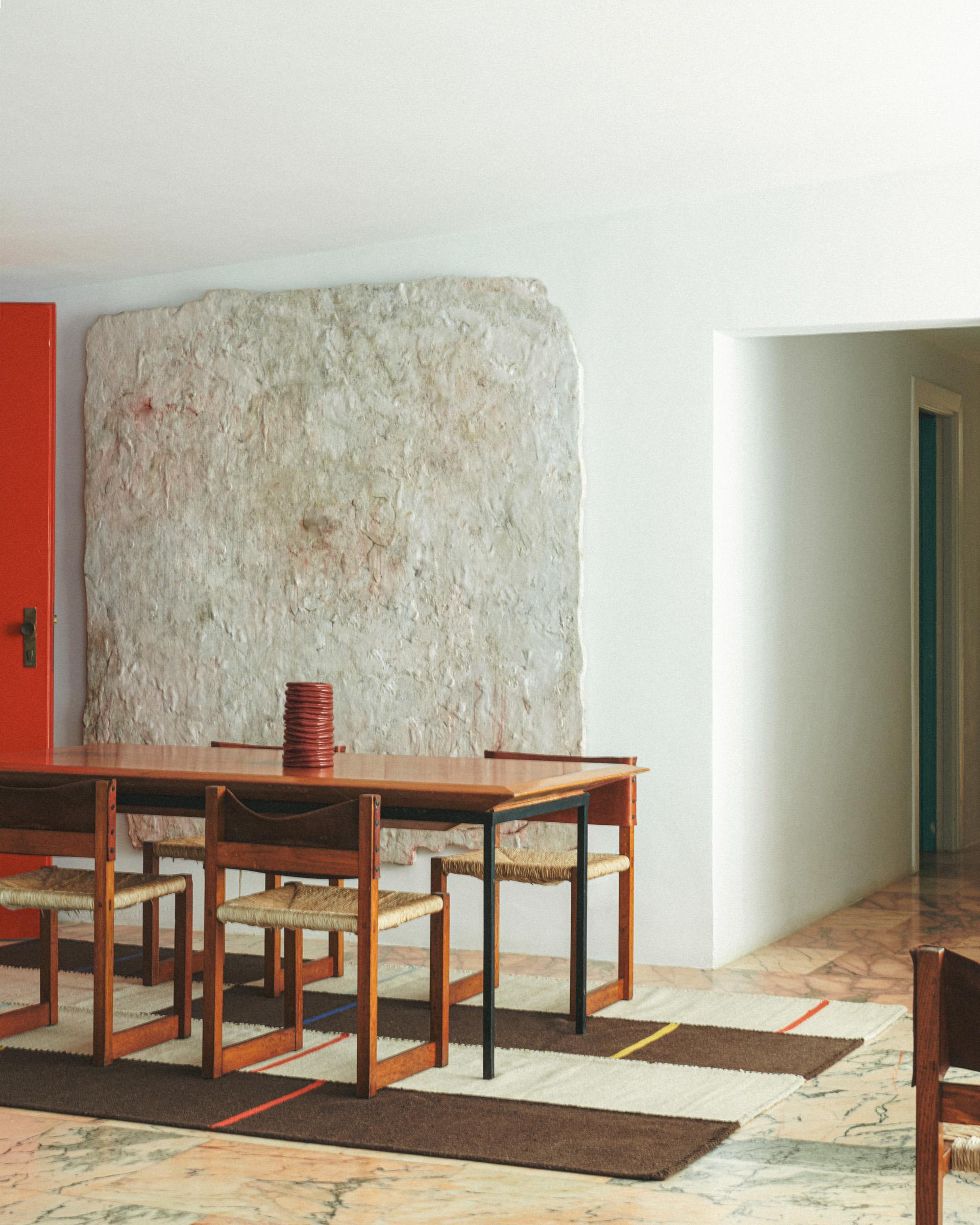 Image of a dining room with a wooden table and chairs and a brown and white flatwoven rug underneath. The back wall has a large stone on it.