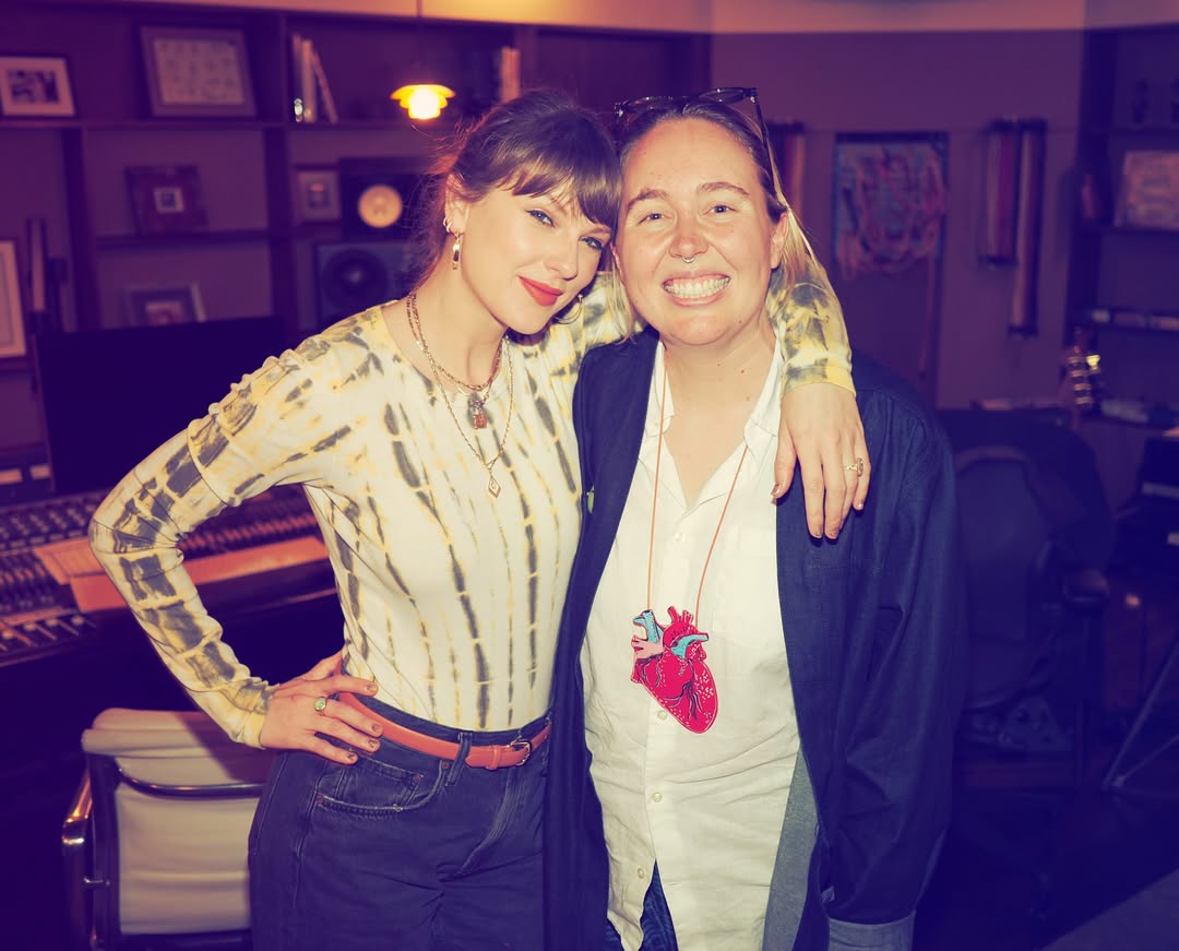 Taylor Swift wearing a tie-dye Agolde bodysuit with black jeans and opal jewelry with brown bangs and a ponytail posing in a recording studio