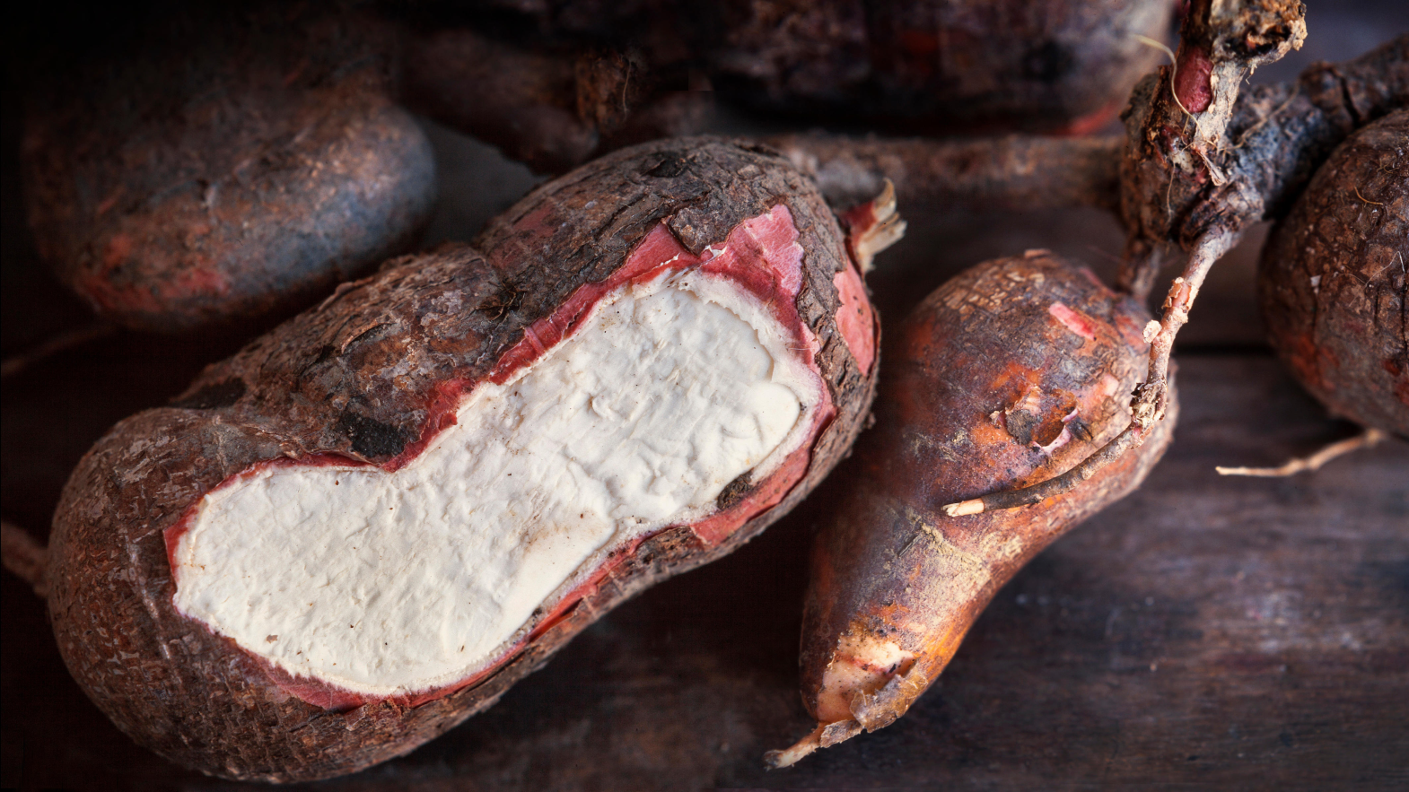 Mai multe rădăcini de manioc se sprijină pe o masă de lemn. Una este tăiată pentru a dezvălui albul din interior.