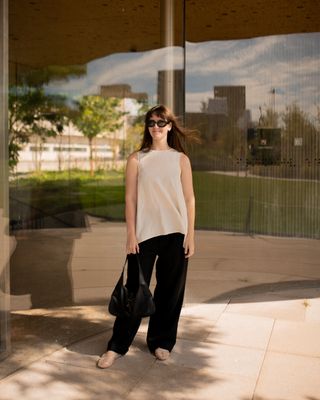 fashion editor Halie LeSavage stands in front of the Copenhagen Opera House wearing a white linen top and black flowy pants with mesh flats and a Gucci purse