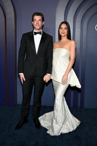 John Mulaney wears a tuxedo while Olivia Munn wears a long white mermaid style strapless dress