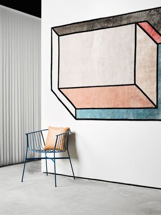 A blue metal chair with an orange-brown leather cushion is set against a wall. There is a geometrical artwork painted on the wall, in pastel colors.