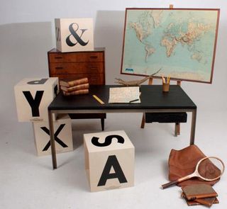 A black table in the foreground with a brown wooden chest of drawers and a map of the world behind it. There are also stacks of large cubes featuring black lettering.