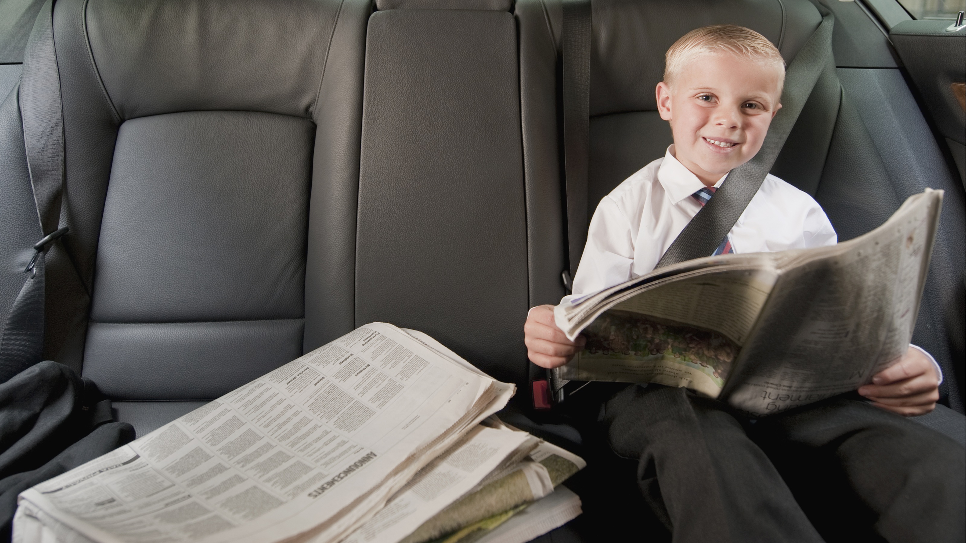 A young boy smiles as he sits in the back seat of a car with a newspaper.
