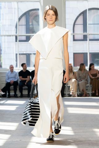 A woman wears a long white dress, black and white sneakers, and a black stripe tote bag.