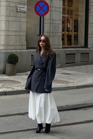 a photo showing what shoes to wear with skirts with a woman standing in the street wearing a gray blazer with a white bubble midi skirt and black knee high boots