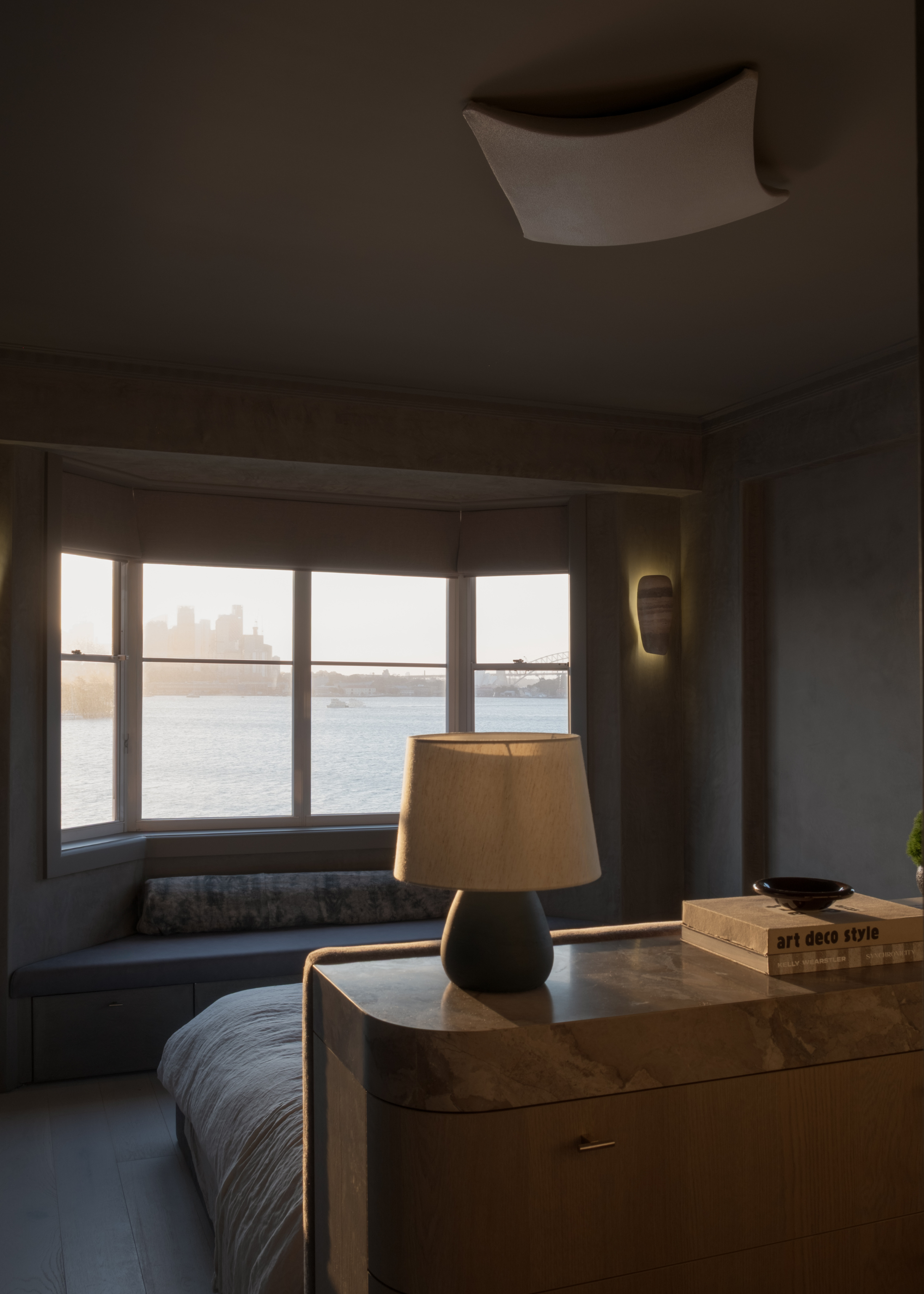 A dark bedroom with a view of a river by a seating nook with a bolster pillow, and a headboard with a small lamp, a stack of books, and a bowl
