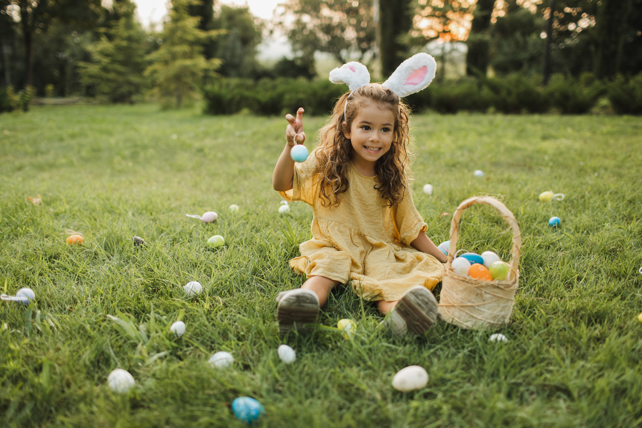 A little girl in a yellow dress wearing bunny ears, smiling while doing an Easter egg hunt.