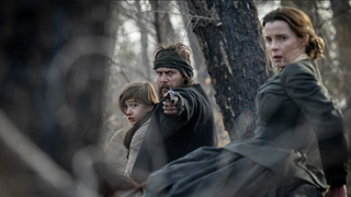 Taylor Kitsch points a fun towards the screen while holding a small boy in American Primeval, while Betty Gilpin looks scared nearby, out in a forest