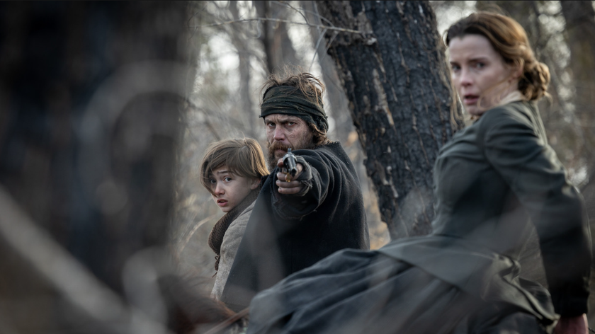 Taylor Kitsch points a fun towards the screen while holding a small boy in American Primeval, while Betty Gilpin looks scared nearby, out in a forest