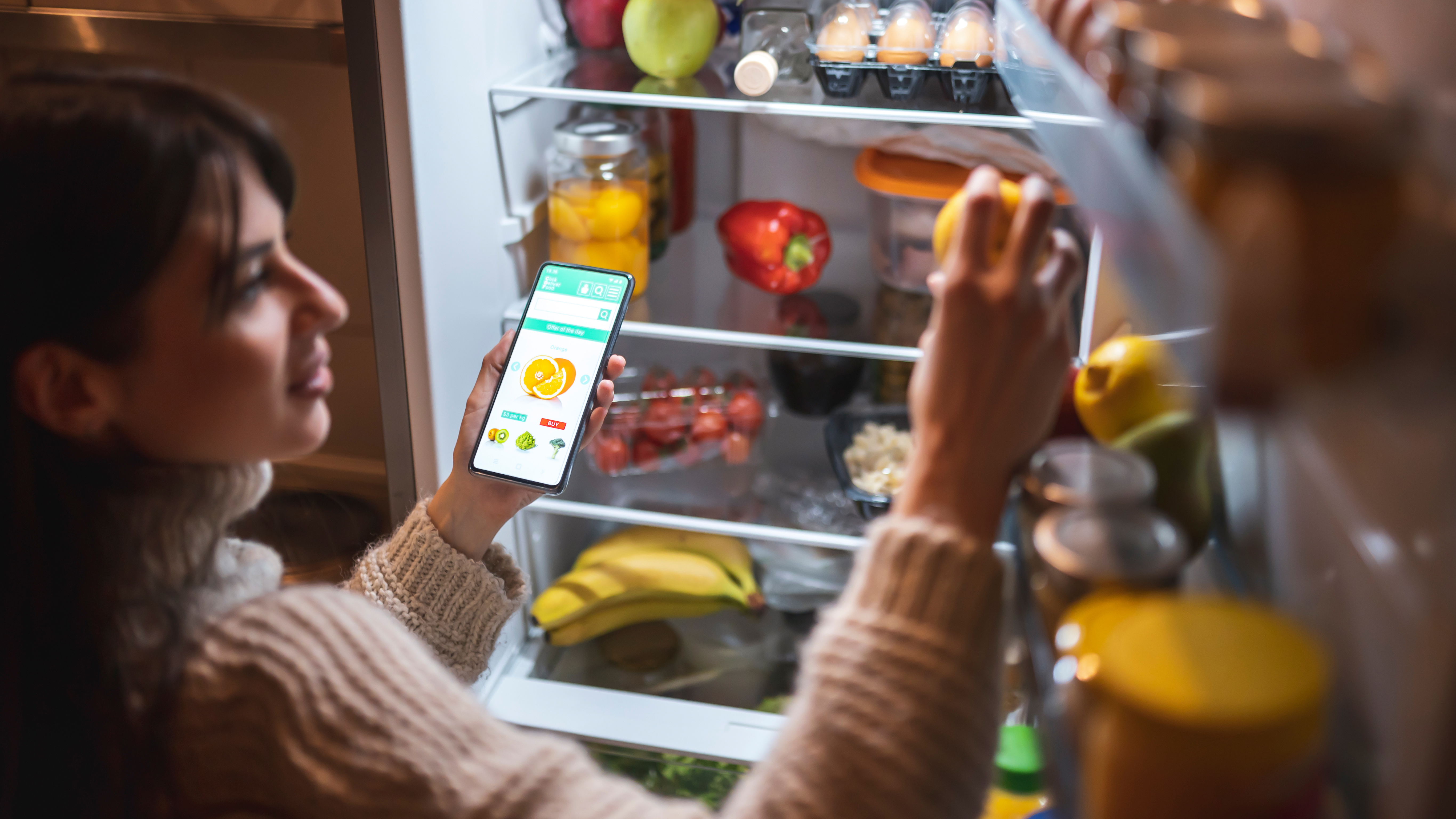 Женщина использует умный холодильник с морозильной камерой с телефоном в руке