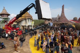 A crowd gathers on a 'Wicked' set styled like Munchkinland and the Yellow Brick Road, with a giant square light hanging above.