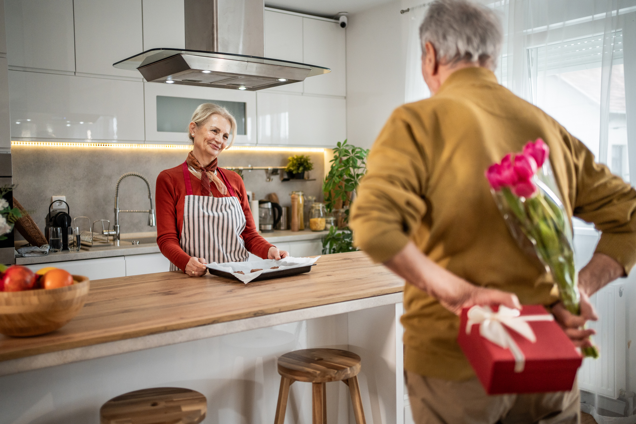 A senior man carries flowers and a gift behind his back to surprise his wife. 