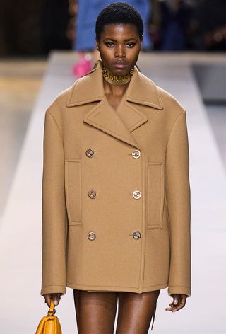 A model on Gucci's fall 2024 runway wearing a tan boxy peacoat styled with a gold choker necklace and a yellow handbag.