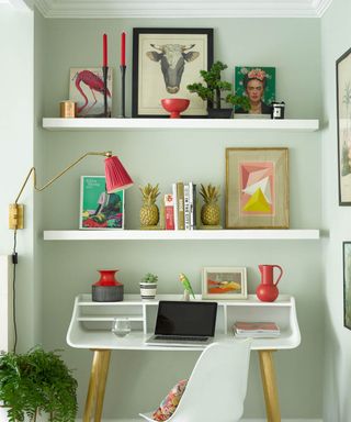 A home office with mint green walls, two white wooden shelves with colorful wall art and books on, a white desk with plants and a laptop on, and a pink and gold lamp over the desk with a spiky green plant below