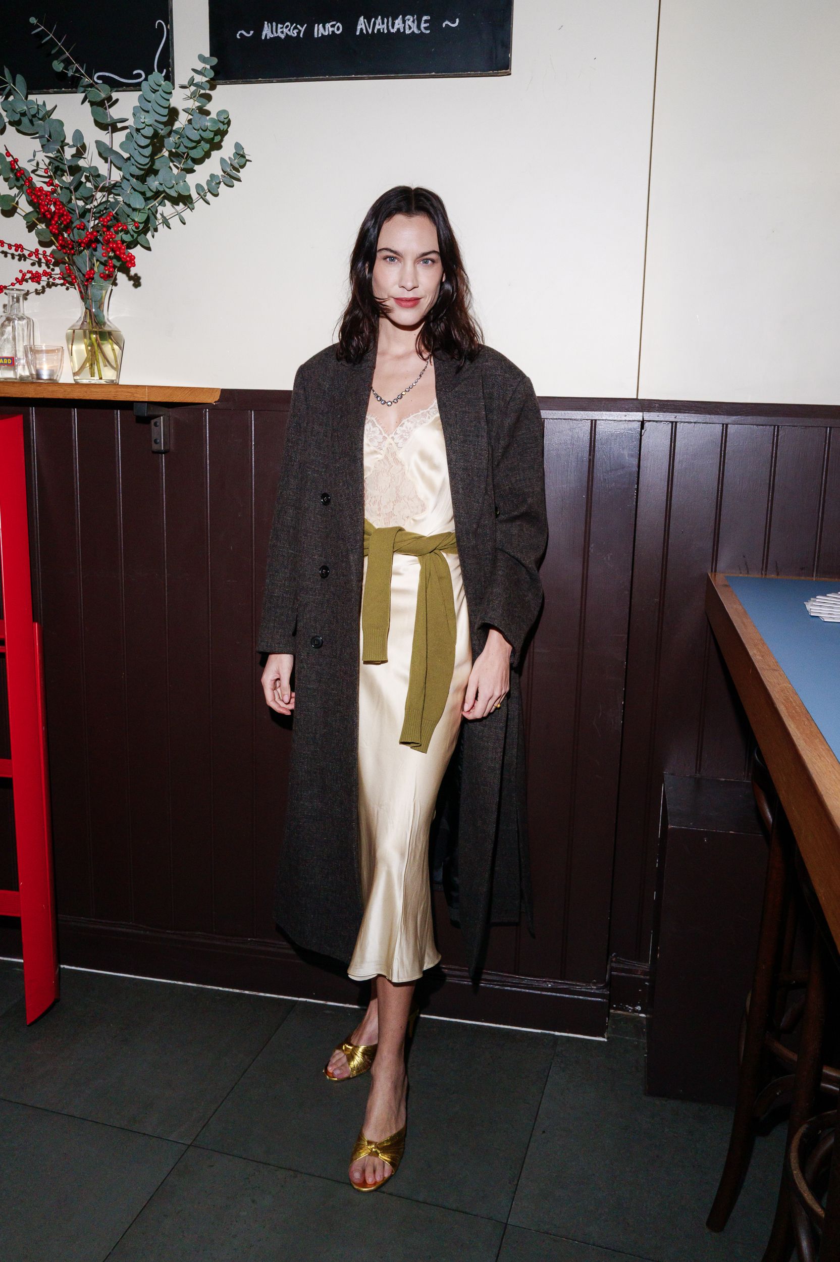 An image of Alexa Chung wearing a Reformation slip dress, grey coat, olive cardigan and gold heels.