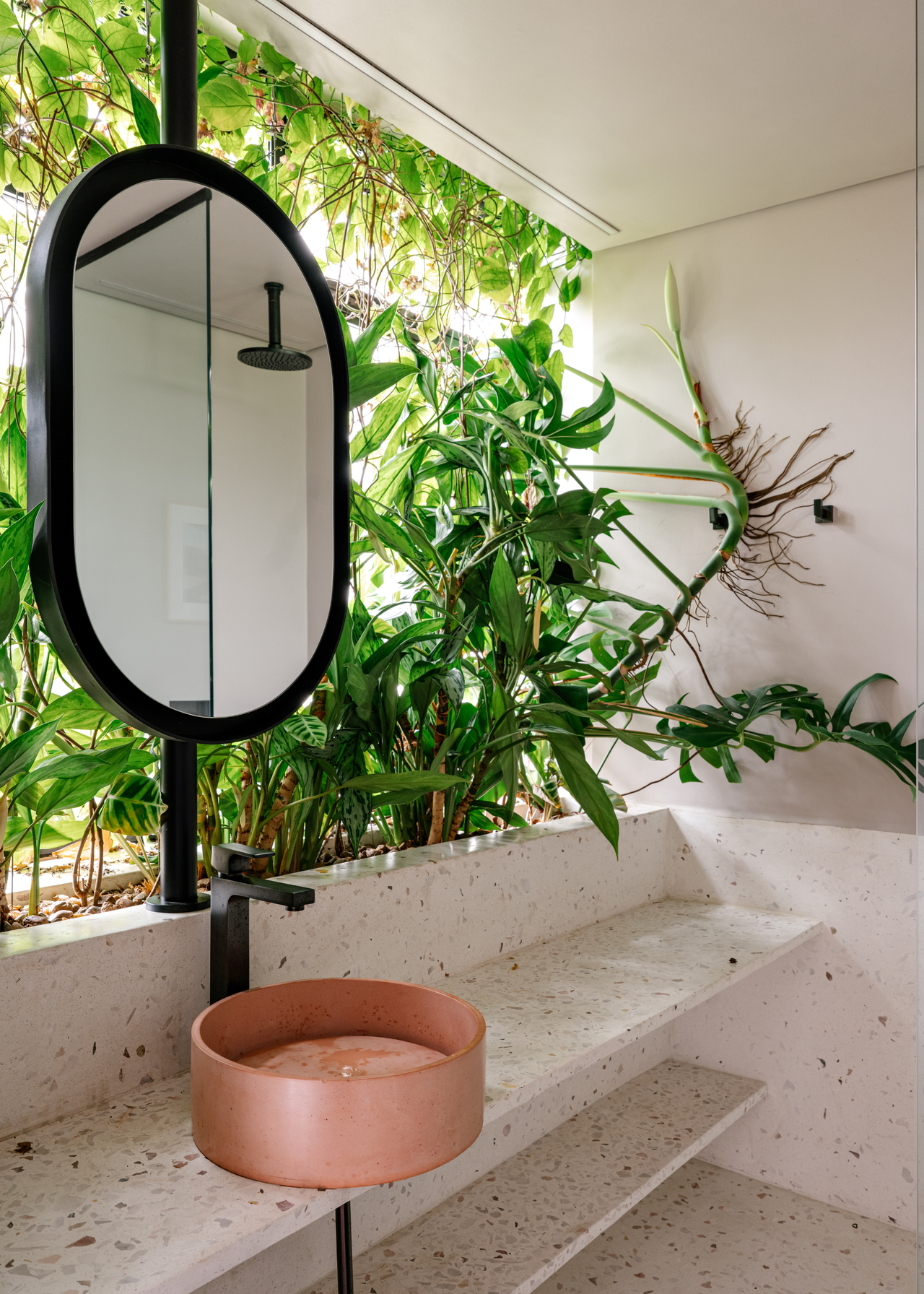 A bathroom with a terrazzo countertop, a small blush sink and an oval mirror backed by an in-built counter garden